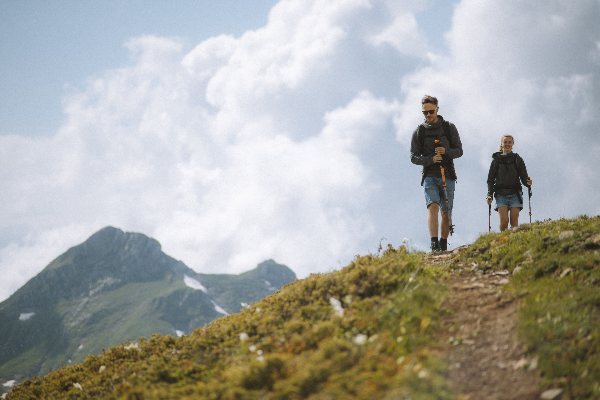 Two hikers walking a mountain ridge trail with poles under a bright sky